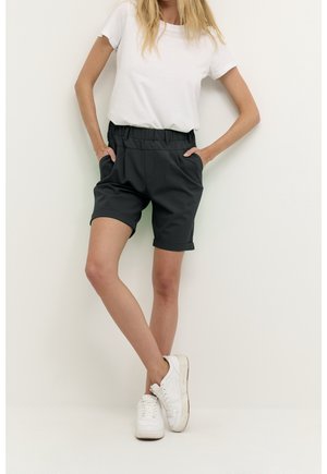 Femme portant un short noir à ourlet roulé, un t-shirt blanc et des baskets blanches, se tenant les mains dans les poches contre un fond blanc uni.