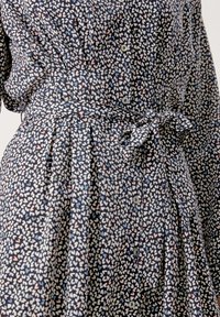 Blumenmuster-Kleid aus dunkelblauem Stoff. Verfügt über Knöpfe, einen Gürtel in der Taille und Falten für zusätzliche Textur und Bewegungsfreiheit.