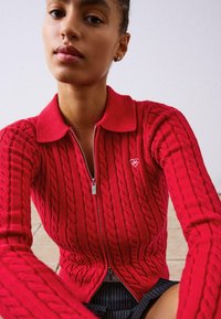 Rdeča zip-up jopica s teksturiranim kabelskim pletenjem in majhnim srčkastim logotipom na levem prsi. Dolgi rokavi s ovratnikom.
