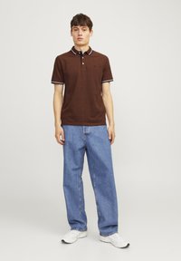 Bruine polo met witte accenten, korte mouwen en drie knopen, gecombineerd met lichtblauwe, losse spijkerbroek en witte sneakers.