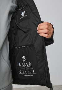 Interieur van een zwarte jas met een technologievak. Het materiaal is glad en glanzend, met merknaam en wasinstructies in witte tekst. Voorzien van een ritsclosure.