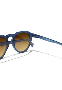 Modré průsvitné sluneční brýle s geometrickým rámem. Čočky mají hnědý přechodný odstín. Ramínka nesou značku a nápis "Vyrobeno ve Španělsku".