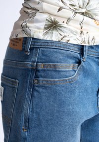 Jean en denim bleu avec une coupe slim, présentant des coutures orange, un patch en cuir à l'arrière et un détail de conception sur la poche.