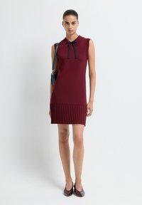 Abito senza maniche color bordeaux con colletto e dettaglio di fiocco blu navy; presenta un orlo plissé nella parte inferiore. La modella tiene una piccola borsa.