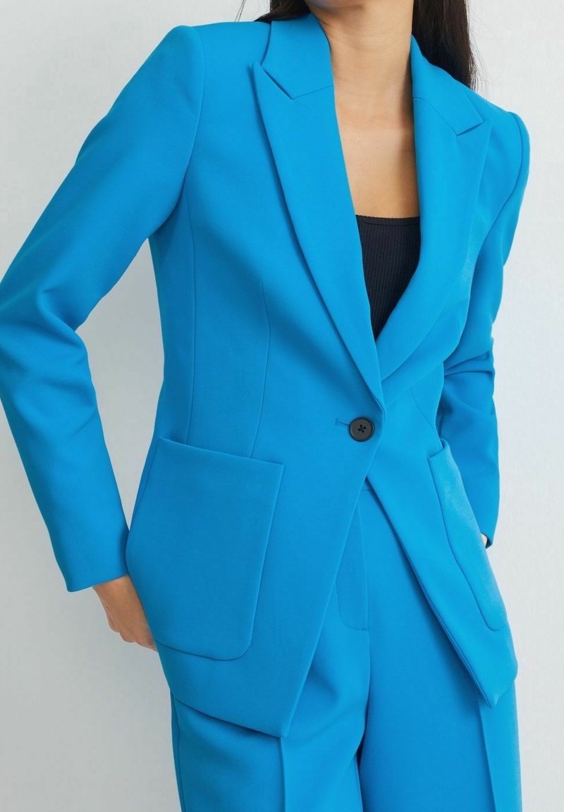 Femme portant un blazer bleu vif sur mesure et un pantalon assorti sur un haut noir, debout avec une main dans la poche.