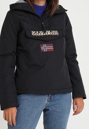 Personne portant une veste noire Napapijri avec un patch du drapeau norvégien et un jean bleu, debout devant un fond uni.