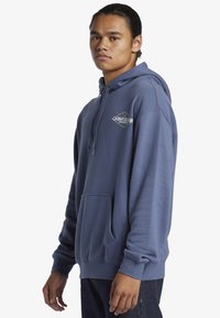 Mikina s kapucňou v tlmenej modrej farbe, s predným vreckom, rebrovanými manžetami a logom na ľavej strane hrude. Mäkka textúra látky.