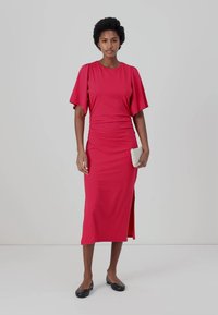 Abito aderente, di lunghezza al ginocchio, di un vivace rosso. Maniche corte fluttuanti, design arricciato in vita e spacco laterale. Abbinato a una piccola clutch leggera.