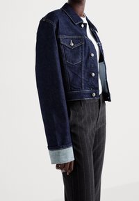 Dark blue denim cropped jacket with button-up front, chest pockets, and light blue cuff accents. Paired with pinstriped trousers.