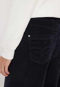 Mörk marinblå kordflöjtbyxor med en bakficka som har ett böljande sömnadsmönster och en metalldekor, i kombination med en vit sweatshirtärm.