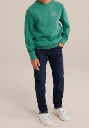 Enfant portant un sweat-shirt vert avec le texte « Mountain Adventure », un jean bleu foncé et des baskets blanches, debout avec une main dans la poche.