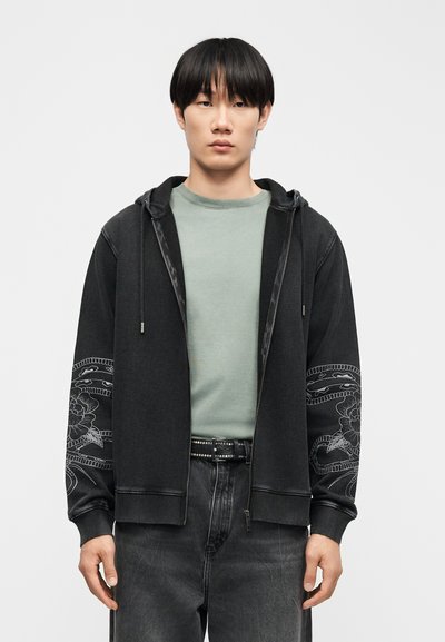 Νέος άνδρας που φοράει μαύρο φούτερ με φερμουάρ, διακοσμημένο με σχέδια λουλουδιών και φιδιών στα μανίκια, ανοιχτοπράσινο πουκάμισο και μαύρο τζιν με ζώνη με τρουκς.