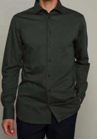 Camicia a maniche lunghe verde scuro in un tessuto testurizzato, con colletto a punta e bottoni scuri lungo il davanti. Vestibilità su misura con orlo arrotondato.