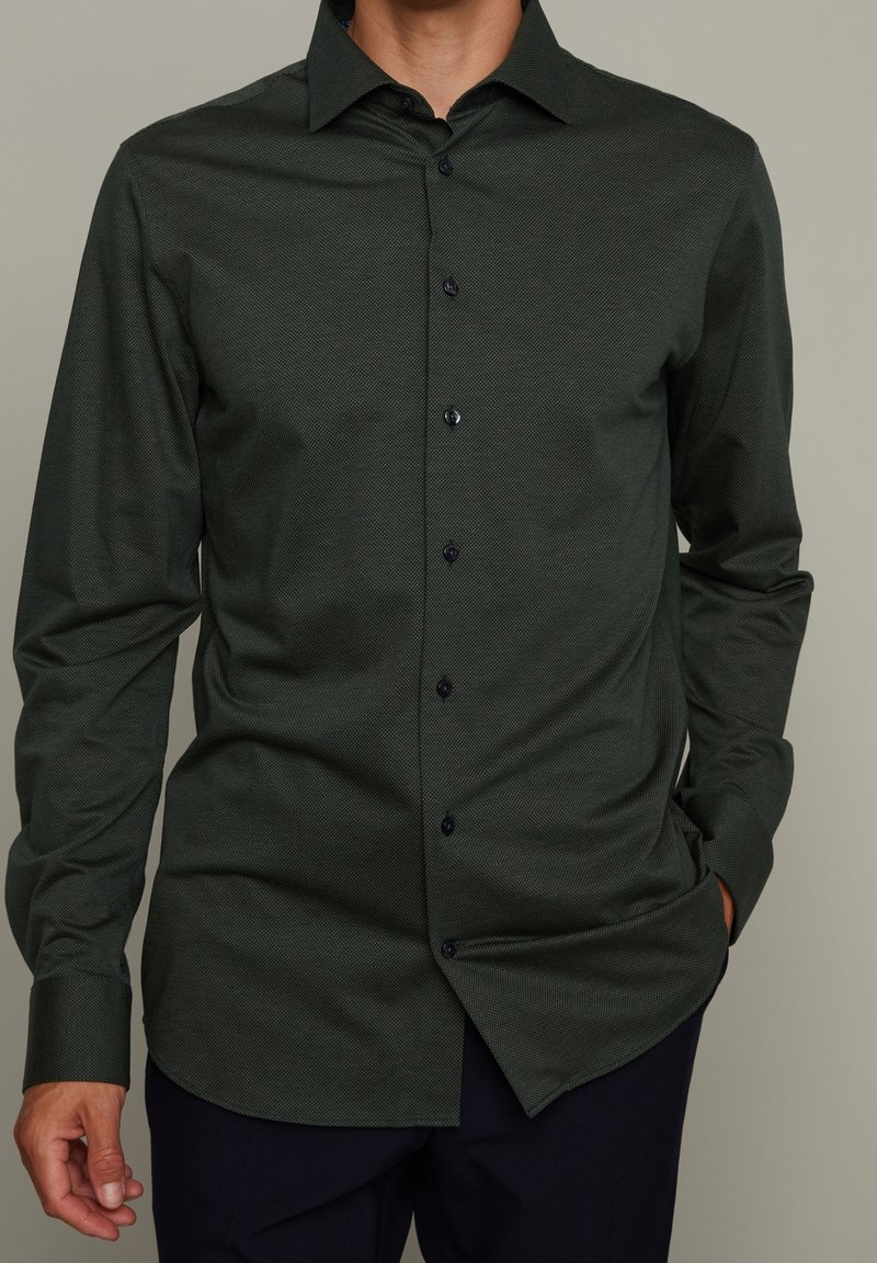 Camicia a maniche lunghe verde scuro in un tessuto testurizzato, con colletto a punta e bottoni scuri lungo il davanti. Vestibilità su misura con orlo arrotondato.