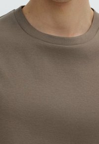 Close-up of a person wearing a plain brown crew-neck shirt, focusing on the neckline and upper chest area.
