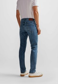 Blå denimjeans med slim fit, fem fickor och en brun logotypbyte på baksidan. Bärs med en vit t-shirt och vita sneakers.