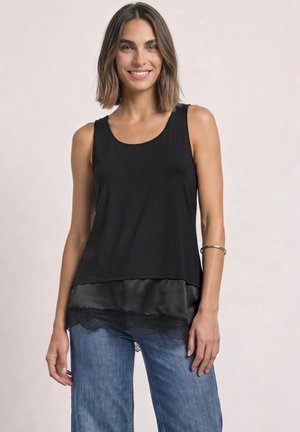 Femme souriante aux cheveux bruns mi-longs, portant un haut noir sans manches avec un ourlet en satin et dentelle, un jean bleu et un bracelet argenté au bras.