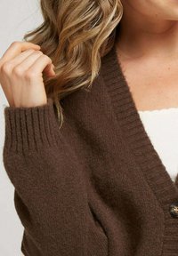 Cardigan en laine marron avec un col et des poignets côtelés, doté d'une fermeture à boutons et d'une texture subtile. Gros plan sur les détails de la manche et de l'épaule.