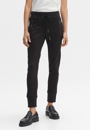 Femme portant un pantalon de jogging noir avec des poches zippées sur les côtés, des mocassins noirs et une chemise blanche rentrée, debout sur un fond blanc uni.