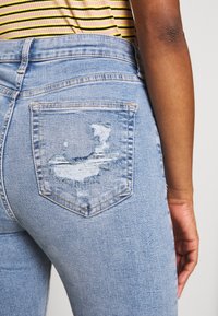 Light blue denim jeans with a distressed back pocket worn by a person in a yellow and black striped top, seen from the back waist down.