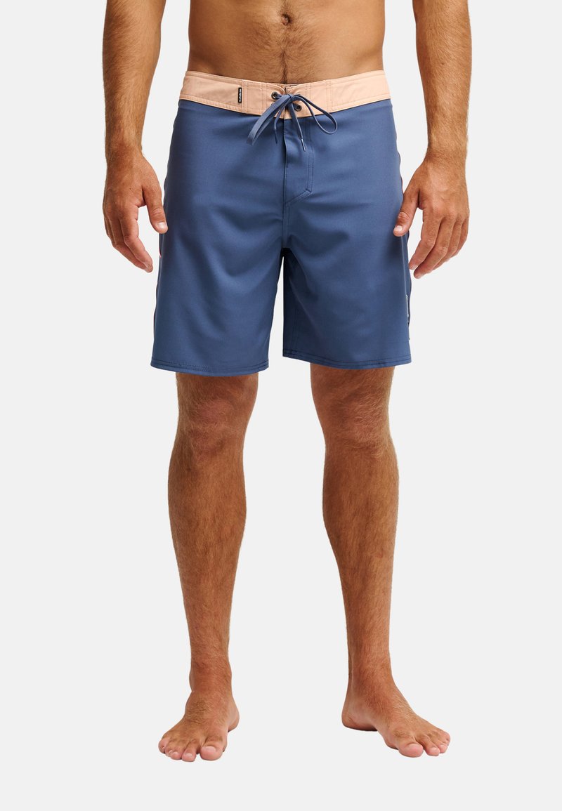 Man wearing blue and beige drawstring swim shorts standing barefoot on a plain white background.