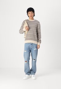 Jersey de punto con un patrón geométrico en beige y marrón, combinado con unos vaqueros ligeros rasgados de color azul y zapatillas blancas, sosteniendo un bolso marrón.