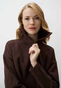 Maglione marrone con zip, colletto alto e tessuto strutturato. Vestibilità ampia con spalle scese; il modello tiene il colletto con una mano.