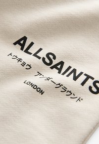 Tissu beige affichant un texte noir avec les mots « ALLSAINTS » et « LONDON », suivi d'un texte japonais dans un style de police similaire.