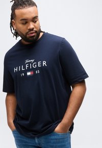 Άνδρας με πλεξούδες, φοράει μια μπλε σκούρα μπλούζα Tommy Hilfiger και μπλε τζιν, με τα χέρια στις τσέπες, κοιτάζοντας προς τα κάτω και προς το πλάι.