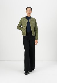 Woman wearing olive green quilted jacket, navy turtleneck, black wide-leg pants, and black shoes, standing against plain white background.