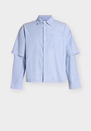 Camicia a righe verticali celeste e bianca con bottoni, maniche corte e lunghe sovrapposte e taschino sul petto su sfondo bianco.