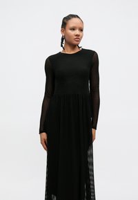 Black long-sleeve dress with textured fabric and a flowing silhouette. Features a fitted bodice and sheer, striped sleeves.