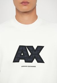 Fehér kereknyakú pulóver, nagy fekete "AX" logóval, amely textúrázott részletezéssel rendelkezik, és alatta az "ARMANI EXCHANGE" felirattal.