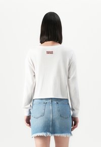 Kvinne med skulderlangt mørkt hår som vender bort, iført en hvit langermet skjorte med teksten "American Vintage" og et slitt denimskjørt.