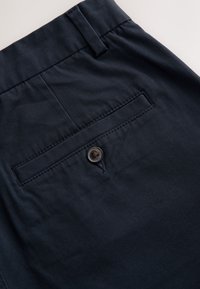 Pantalon bleu marine en gros plan montrant une poche arrière avec fermeture par bouton et une boucle de ceinture sur le bord supérieur.