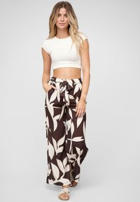 Femme portant un haut blanc court à manches courtes et un pantalon large taille haute avec un grand motif de feuilles blanches sur fond sombre.