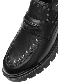 Mocassin en cuir noir avec des clous en métal le long de l'orteil et du dessus, présentant un détail de sangle et une semelle en caoutchouc épaisse.