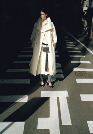 Femme en long manteau crème et jupe, debout sur un passage piéton ensoleillé, les mains dans les poches du manteau, portant des talons noirs et des chaussettes grises.