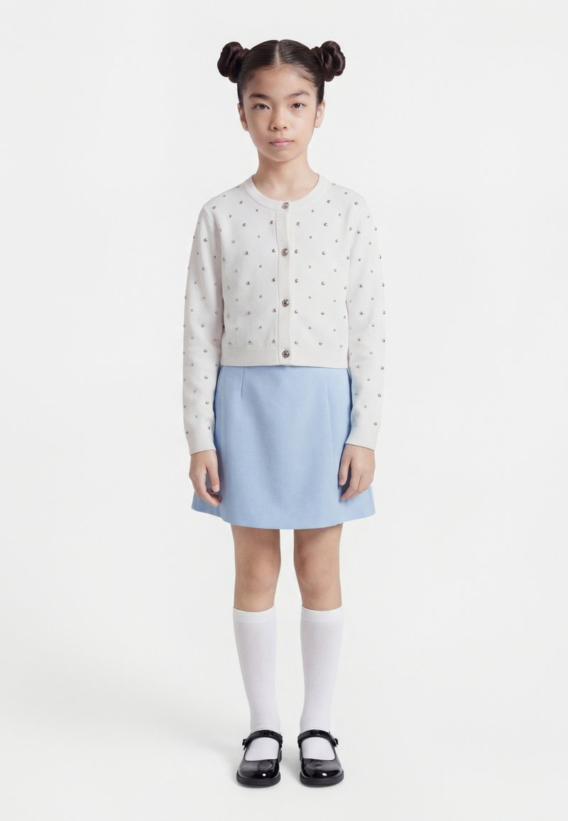 Νεαρό κορίτσι με χτένισμα διπλού κότσου, φοράει λευκή ζακέτα με κουμπιά και μικρά σχέδια, φούστα ανοιχτού γαλάζιου χρώματος, λευκές κάλτσες μέχρι το γόνατο και μαύρα παπούτσια.