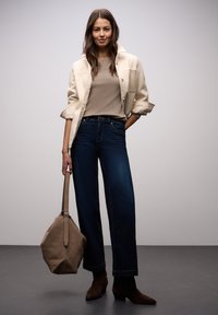 Beżowy top z krótkim rękawem pod rozpiętą kremową kurtką; do tego ciemnoniebieskie jeansy o szerokich nogawkach i brązowe zamszowe botki; trzyma jasnobrązową torbę.