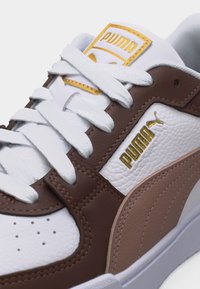 Κοντινή λήψη λευκού και καφέ αθλητικού παπουτσιού Puma με λευκά κορδόνια και χρυσά λογότυπα Puma στη γλώσσα και στη πλευρική επιφάνεια.
