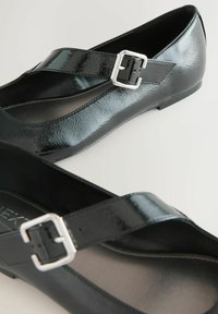 Fekete lakkbőr balerina cipő keresztpánttal és ezüst csattal. Simás felületű, kerek orrú, alacsony sarkú.