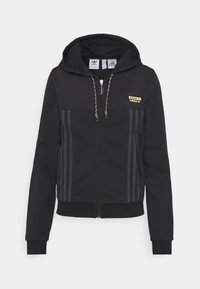 Giacca nera con cappuccio e zip, realizzata in tessuto morbido, caratterizzata da strisce a contrasto, un patch con logo dorato e polsini e orlo a costine.