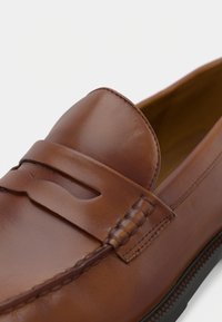 Mocassin en cuir marron avec un dessus surpiqué et légèrement surélevé, orné d'un détail découpé décoratif. Texture lisse et bout arrondi. Semelle en caoutchouc avec un motif de traction.