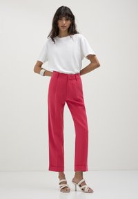 Donna con una camicia bianca a maniche corte, pantaloni fucsia a vita alta, sandali con tacco bianchi e un braccialetto bianco, in piedi con le mani sui fianchi.