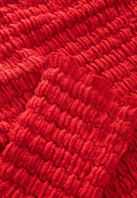 Tissu rouge texturé avec une apparence légère et duveteuse, présentant une série de motifs arrondis en relief et un détail de bord ondulé.