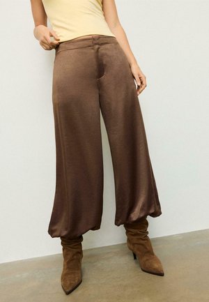 Kobieta w błyszczących, brązowych, szerokich spodniach włożonych do luźnych brązowych zamszowych kozaków na obcasie, stojąca przy gładkiej jasnej ścianie.