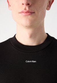 Suéter negro de algodón con un cuello acanalado y un logotipo blanco de Calvin Klein en el pecho, que enfatiza un diseño simple y limpio.