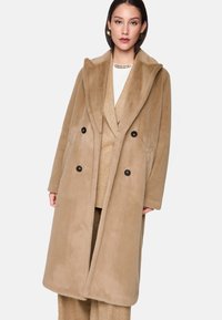 Beige dubbelzijdige jas met een brede kraag, gemaakt van zachte stof. Voorzien van zwarte knopen en een ontspannen pasvorm, gelaagd over een gouden outfit.