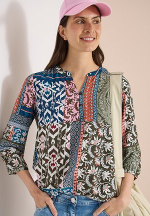 Uśmiechnięta kobieta ubrana w patchworkową bluzkę z długim rękawem, różową czapkę, niebieskie dżinsy i nosząca beżową torbę na ramię.